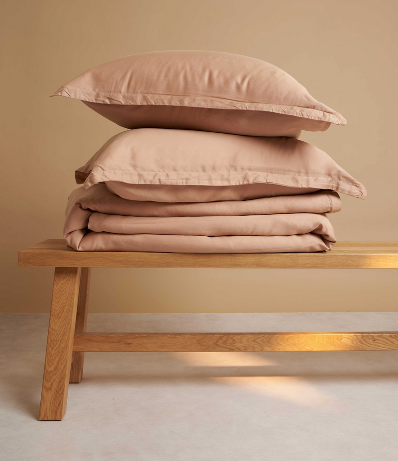 Een gestapeld setje kussens en beddengoed op een houten bedbankje, dienend als navigatielink naar de slapen collectie met subcategorieën zoals bedbankjes, dekbedden, dekbedovertrekken, hoeslakens, kamerschermen, kussenslopen en nachtkastjes.