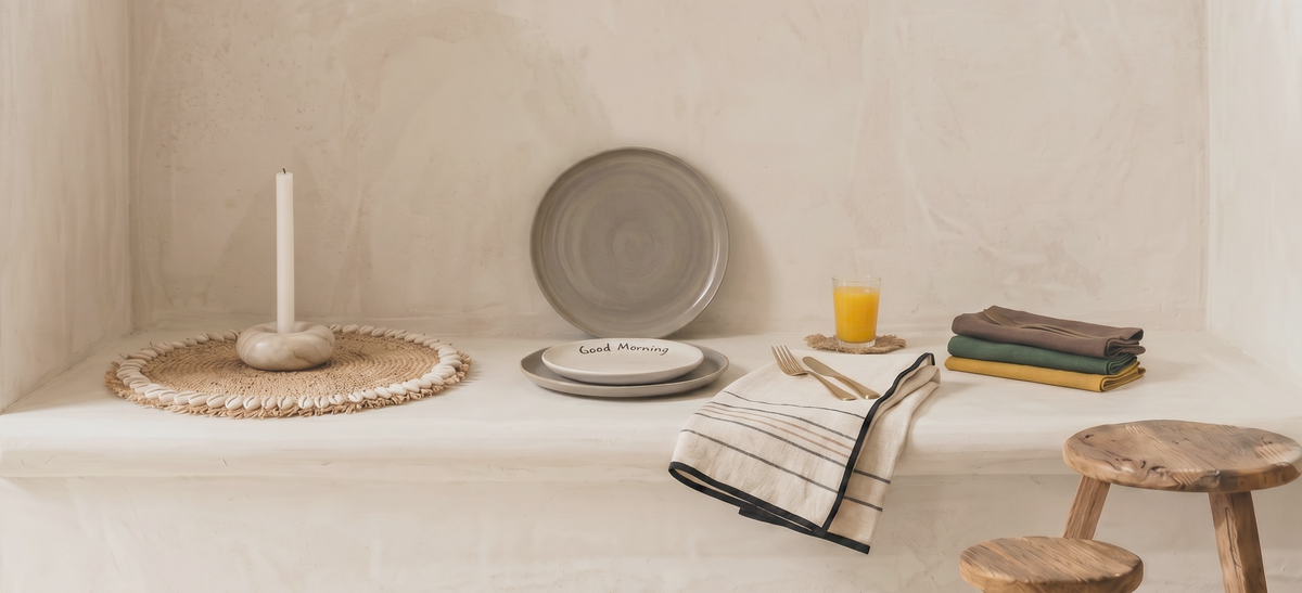 een sfeervolle, brede banner-afbeelding voor de veelgestelde vragen pagina, met een rustieke, ingebouwde lichte cementen bank gestyled met handgemaakt servies, een 'Good Morning' ontbijtbord, een glas jus d'orange, linnen servetten en een minimalistische kandelaar binnenshuis